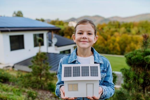 Mädchen hält Modellhaus mit Photovoltaikanlage auf dem Dach vor einem richtigen Haus mit Photovoltaikanlage
