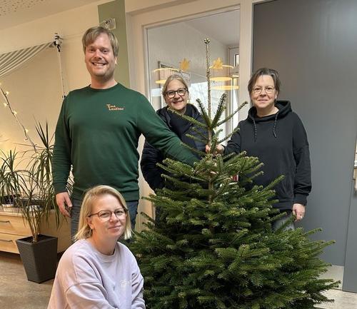 Herr Fichter hält einen Tannenbaum, den er gerade in den Kindergarten liefert. Eine Erzieherin, die Kitaleiterin und Frau Stiller von den Stadtwerken stehen neben dem Baum.